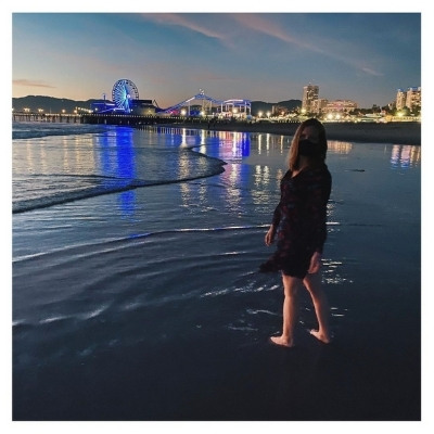 Bryn Donovan barefoot on the beach near Santa Monica pier at twilight, wearing mask, with illuminated Ferris wheel and other city lights in background