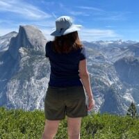 BRYN DONOVAN, back turned, wearing shorts, a T-shirt, and a sun hat. In the background: tall mountains of Yosemite National Park, blue sky