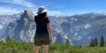 BRYN DONOVAN, back turned, wearing shorts, a T-shirt, and a sun hat. In the background: tall mountains of Yosemite National Park, blue sky