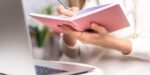 close up of woman's hands as she writes in pink journal; laptop in foreground