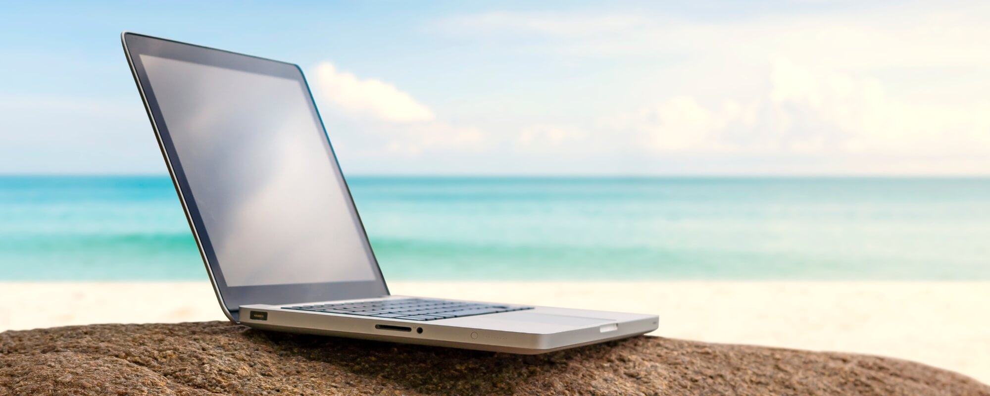 laptop on rock with beach and blue sky in background