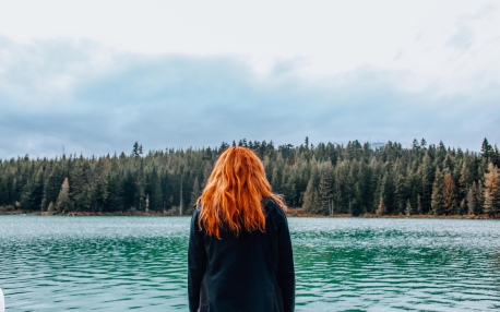 redhead woman from behind, looking at lake, pines, and sky