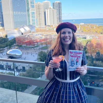 author Bryn Donovan standing on the balcony at Cindy's Rooftop in Chicago, holding her book HER KNIGHT AT THE MUSEUM and a Cosmopolitan. Chicago's iconic Bean sculpture, skyscrapers, and Lake Michigan are in the background.