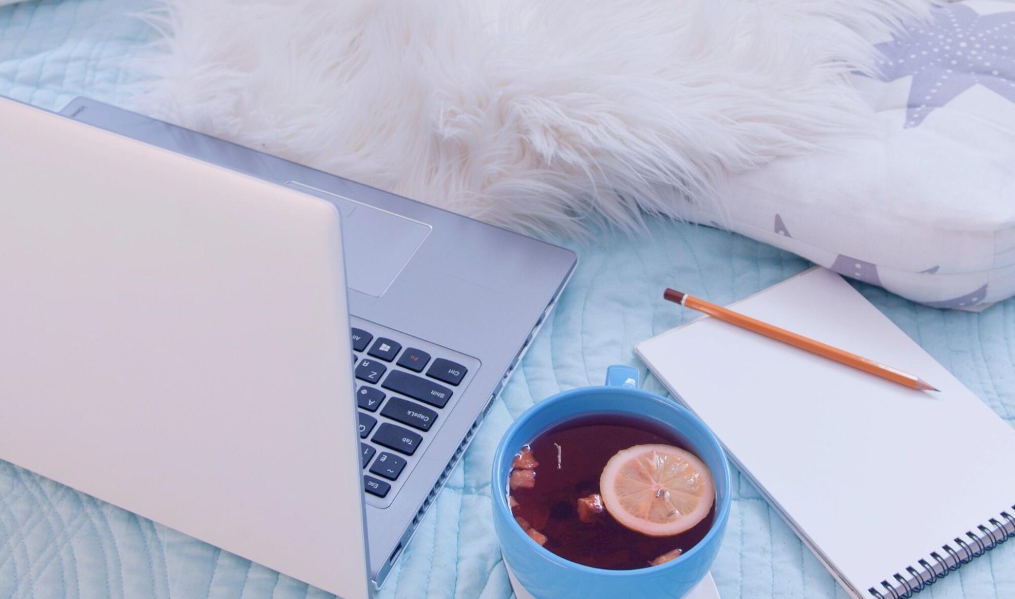 laptop, mug of tea, notepad, pencil, and furry throw on bed