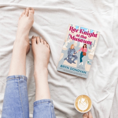 woman's bare feet, a latte, and the romcom book Her Knight at the Museum on a bed